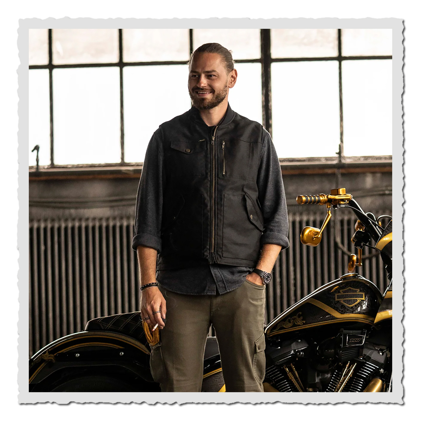 Mann in schwarzer Weste steht in Werkstatt neben Motorrad, große Fenster im Hintergrund, dunkle Kleidung, goldene Details am Bike.